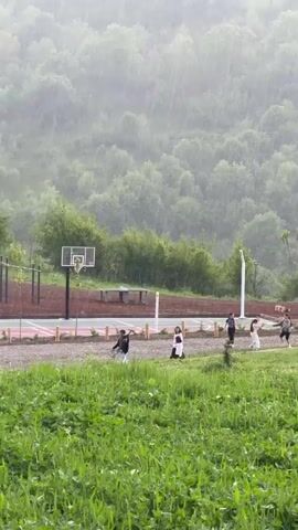 The video shows a scene in what appears to be a rural or mountainous area during a rainy day. The camera is in motion, moving past a sports area with a basketball court and running track. Several people are seen walking or running along the track, though their details are not clear due to the rain and distance. In the background, there's a hillside covered in dense, green trees, shrouded in mist or rain. The camera continues to move, passing a long, modern building with light-colored walls and a flat roof, situated on a grassy lawn. The overall environment appears wet and overcast due to the rainy conditions.