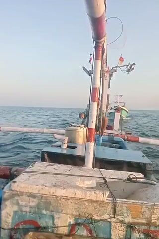 A first-person perspective video shows the deck of a fishing boat on the ocean. The boat is weathered and worn, with peeling paint and some rust visible. A tall, striped pole with red and white bands stands prominently in the center of the frame. Further back on the boat, two men are visible. One man, wearing a blue shirt, is seated and appears to be working. Another man, wearing a light blue shirt and dark pants, is standing and looking out at the sea. A second, smaller fishing boat is visible in the distance, also on the water. The sky is clear and a pale blue, indicating it is likely daytime. The ocean is calm with small waves.
