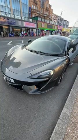 A gray McLaren sports car is parked on a street next to a curb. The car's distinctive butterfly doors are open. Behind the car, across the street, there are shops with colorful signs and a few pedestrians walking. The car is the primary subject of the video.