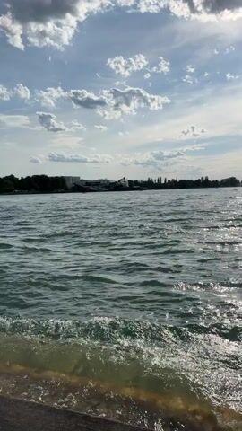 A video shows a body of water with small waves lapping against a shore. The sky above is partly cloudy with the sun peeking through the clouds. A distant shoreline with buildings and trees is visible across the water. The video pans slightly to the left, showing more of the water and sky.