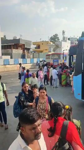 The video captures a busy scene at a bus stop or terminal in Chikkaballapura, India, during daylight hours with a clear sky. A large number of people, including men, women, and children of various ages, are present.

The primary activity involves people queuing, walking, and boarding buses. On the right side of the frame, a blue and black bus is actively loading passengers, with several individuals seen entering through its open door. In the background, another red and white bus is also parked, suggesting it's also at a stop, with more people gathered around it. Individuals are seen carrying bags and backpacks, indicative of travel.

The setting features various buildings in the background, some appearing residential while others might be commercial or industrial. Utility poles and power lines are visible against the sky. The overall atmosphere is one of movement and typical public transportation activity. No specific text on the buses or signs is clearly legible.