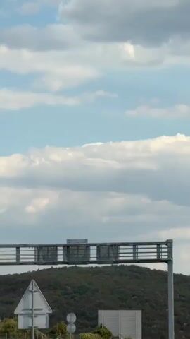 The video captures a view from a moving vehicle on a highway. In the foreground, a highway sign structure with horizontal bars is visible, along with a vertical support pole. To the right, a concrete wall lines the road, and a speed limit sign indicating 130 is present. Further down the road, other vehicles can be seen, including a dark-colored car. The landscape features rolling green hills covered in vegetation. In the background, mountains are visible with some structures and antennas. The sky is partly cloudy, with white clouds against a blue backdrop. A digital display on the highway sign shows the temperature as 32.1°C. Traffic lights are also visible in the distance.