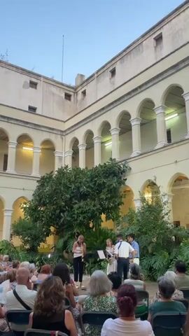 The video shows a courtyard performance in front of an audience. The courtyard is enclosed by a multi-story building with arched windows and walkways. Lush green plants and a large tree are visible in the center of the courtyard. Four musicians are performing on a raised platform. One female musician is standing and playing a flute. Behind her, three other musicians are playing instruments. Two appear to be playing saxophones, and one is playing a guitar. The audience is seated in chairs facing the performers. The lighting suggests it is either early morning or late evening. The overall scene depicts an outdoor concert in an architectural setting.