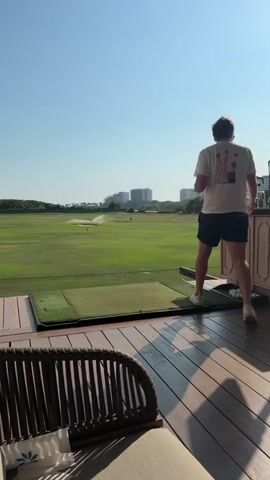 The video shows a man at a golf driving range. He stands at a small wooden cabinet containing golf clubs and balls, overlooking a large, green golf field being watered by sprinklers. In the background are buildings and a clear blue sky. The man is wearing a white t-shirt, dark shorts, and light-colored shoes. He appears to be selecting a golf club from the cabinet. The camera then shifts focus to a nearby area with wooden tables and chairs, where a glass of amber-colored liquid is sitting on one of the tables. Another man wearing red patterned shorts walks by.