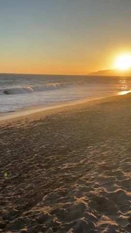The video shows a beach at sunset. The sun is setting over the ocean, casting a warm orange glow across the scene. Waves are gently breaking on the shore, and the sand is covered in footprints. A single person is walking on the beach in the distance. The sky is clear and a gradient of blue and orange. There is a person in the distance walking along the shore.