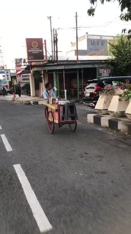 The video shows a street scene in Sidoarjo, East Java. A man is pushing a food cart along the side of a road. The cart is red and yellow, with a cooler and a stool. The man is wearing a blue shirt and a white hat. He appears to be selling food or drinks.

The scene is set during the day, with sunlight casting shadows. The weather appears to be warm and clear. The road is paved and has white lines marking the lanes. There are buildings on either side of the road, including shops and houses. There are also trees and plants visible in the background.

Notable details include the text "THE NUSANTARA" visible on a sign in the background. The overall mood of the scene is one of everyday life and commerce in a Southeast Asian city. The man's expression is neutral, and the activity seems routine.