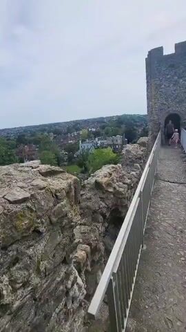 The video shows the interior and exterior of a castle.  The camera moves from a stone wall with a metal handrail along the edge of the castle toward a stone tower and then turns to look over the edge and into the castle's courtyard. People are walking around. The castle is made of stone. The castle walls are old and weathered. Outside the castle, there are trees and buildings in the distance. The sky is overcast.