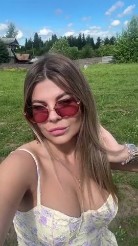 A young woman with long blonde hair and pink sunglasses is taking a selfie outdoors. She is wearing a yellow dress with a floral pattern. She is smiling and her hair is blowing in the wind. Behind her is a grassy field with trees and a fence. In some shots, a car is visible.