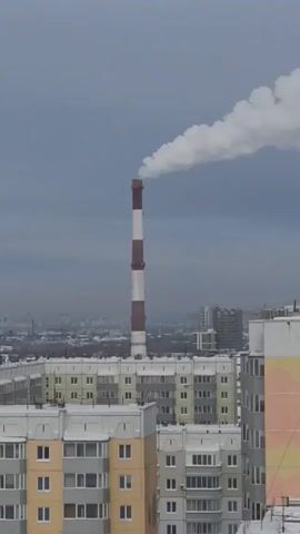 The video shows a tall industrial chimney billowing white smoke into a gray, overcast sky. The chimney is painted with alternating red and white stripes. In the foreground, there are several apartment buildings with a mix of yellow, gray, and peach-colored facades. Some buildings have balconies. The rooftops are dusted with snow. In the background, the skyline of a city is visible through a hazy or smoggy atmosphere. There's a construction crane visible amongst the buildings in the distance. The overall scene suggests a cold, urban environment with industrial activity.