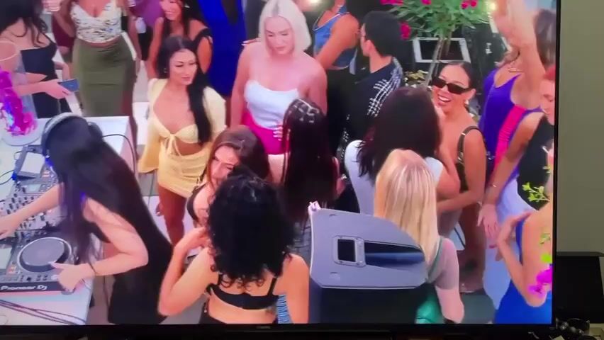 The video shows a group of people at what appears to be a party. A DJ with long, dark hair and headphones is operating a Pioneer DJ mixer. Many of the attendees are women, dressed in various outfits, some revealing. There are also a few men present. Some people are holding drinks. The atmosphere seems lively, with people dancing and interacting. The party seems to be taking place outdoors, possibly on a patio or rooftop, with plants visible in the background. The video is being viewed on a Xiaomi TV.