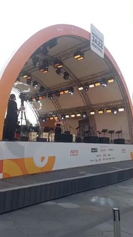 A wide shot shows an outdoor stage at the Moscow Jazz Festival. The stage is covered by an orange arched canopy. A band is performing on stage, with a female singer at the microphone in the center. A cameraman is filming the performance from the left side of the stage. The stage is decorated with the festival logo and sponsors' names. The ground in front of the stage is paved with grey tiles. The weather appears to be overcast.