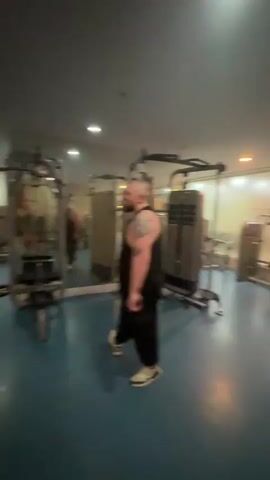 The video shows the interior of a gym. There are several weight machines, benches, and free weights on the blue floor. Several individuals are present, some wearing workout attire. One man in a black tank top and black pants is walking towards the right of the frame. A man in an olive green tank top and shorts is standing in front of a Smith machine, presumably taking a photo or video of himself. Music plays in the background.