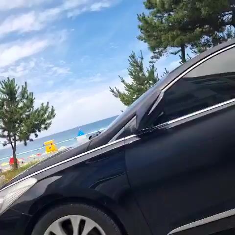 The video shows a view of a beach from the perspective of someone in or near a car. The camera pans across a parking area with at least two cars, one black and one white. Beyond the cars, there is a wooden walkway leading to a beach. The beach is dotted with colorful umbrellas and tents. The ocean is visible in the background, and the sky is blue with some clouds. Pine trees are scattered along the edge of the beach. The video is short and does not contain any discernible people or specific actions.