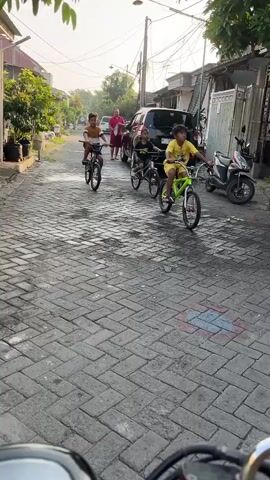 The video shows a group of children riding bicycles down a paved street. The street appears to be in a residential area with houses on either side. There are several parked vehicles, including a car and a motorcycle. The children are of various ages and are all wearing casual clothing. They appear to be enjoying themselves as they ride their bikes. The sun is shining, suggesting it is daytime.