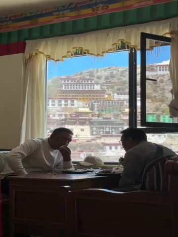 Two men are sitting at a wooden table, eating in front of a large open window. The man on the left is wearing a white long-sleeved shirt, and the man on the right wears a gray shirt and eyeglasses. He is using chopsticks. The table has two plates of food, a hat, and some utensils. Behind them, the window shows a view of a large, multi-tiered complex built on a mountainside under a bright blue sky. The room is decorated with colorful patterns along the ceiling. The window is framed by white curtains with gold trim.