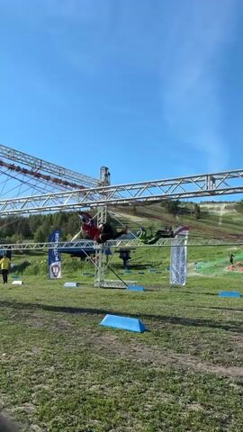 A person is traversing a high obstacle course, suspended from a metal structure with cables and ropes. The course is set in a grassy field with blue markers on the ground. In the background, there's a hill with trees, and a small building. A person in a yellow jacket is walking on the ground below. Banners with text in Russian are visible beside the course. The sky is clear and blue. Someone shouts encouragement in Russian.