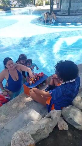 In a pool area, several people are visible. In the foreground, a woman in a blue swimsuit stands in the water near the edge, reaching towards a game board with small figures on it. To her left, another woman holds a child. A person with dark curly hair wearing an orange and blue shirt is seated on rocks by the pool. In the background, other individuals are in the pool, and palm trees and buildings surround the area. A fountain sprays water in the middle of the pool. The overall scene appears to be a recreational setting.
