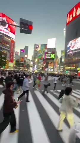 The video shows a busy intersection in Tokyo, Japan, likely Shibuya Crossing, at what appears to be dusk or early evening. Numerous pedestrians are walking across the zebra-striped crosswalks in various directions. Overhead, bright billboards and large screens display advertisements. The sky is light and the overall atmosphere is energetic and bustling.  Individuals are wearing a mix of casual clothing.  Traffic lights are visible, and the overall setting is a vibrant urban environment.