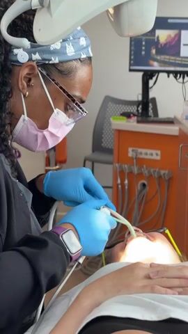A dentist, wearing blue gloves, a pink mask, and a headband with a cat pattern, uses dental tools to examine a patient. The patient is lying down, with their mouth open, illuminated by a dental lamp. In the background, there is an orange cabinet with various dental instruments and a computer monitor displaying an image.