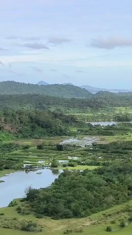 The video shows a scenic aerial view of a lush green landscape. The landscape includes rolling hills covered in dense trees, bodies of water that appear to be lakes or flooded fields, and a coastline with a beach and the ocean. The sky is partly cloudy with a light blue hue. The camera pans slowly across the landscape, offering a wide panorama of the natural environment. The video appears to be a drone shot or from a high vantage point, capturing the expansive beauty of the area.