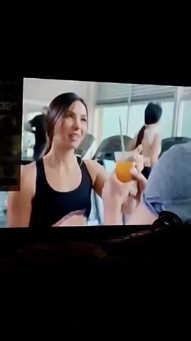 The video depicts an indoor scene set in what appears to be a gym or fitness center, illuminated by daylight streaming through large windows. A woman with long dark hair, wearing a black tank top, is holding a glass with an orange liquid (possibly juice) and a smartphone. She is smiling and looking at a man with dark hair, who is wearing a grey t-shirt. The man is also smiling and appears to be speaking to her. In the background, other individuals are visible, including a woman with red hair, and several treadmills, suggesting ongoing exercise activities. The interaction between the main woman and man seems friendly and positive. On the far left of the frame, a partially obscured screen displays the text "32'". Spanish dialogue is audible, with the man saying, "Te veías tan hermosa..." (You looked so beautiful...).