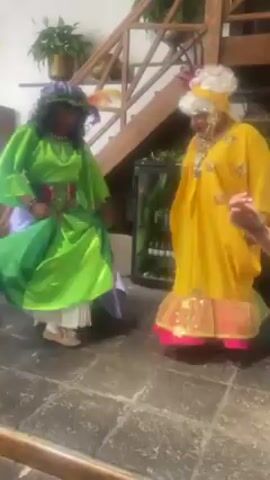 Two people in drag are dancing on a stone tile floor. The person on the left is wearing a green, purple and white dress with a colorful belt and a floral headpiece. The person on the right is wearing a yellow dress with pink trim and a white wig. There is a wooden staircase and potted plants in the background. A refrigerator with bottles inside is visible in the background as well.