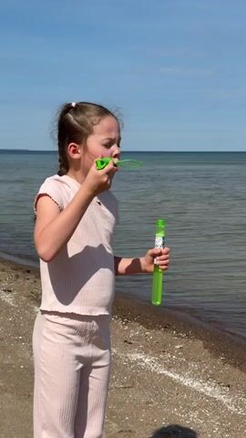 A young girl stands on a beach, blowing bubbles. She has light brown hair in two braids and is wearing a pink, striped, two-piece outfit. She holds a green bubble wand and a container of bubble solution. Behind her, the water stretches to the horizon under a clear blue sky. The beach is sandy with small white shells scattered along the shoreline.
