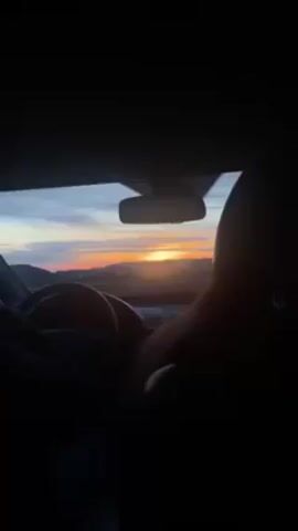 The interior of a car is seen from the backseat. The front seats are visible in silhouette. The driver is holding the steering wheel, and a passenger is in the passenger seat. Through the windshield, a sunset is visible over a hilly landscape. The sky shows hues of orange, pink, and blue. A rear-view mirror hangs from the car's ceiling.