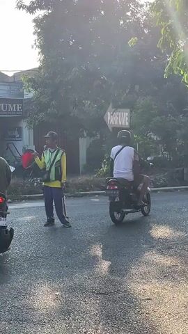 A man in a yellow vest and blue pants stands in the middle of a street, holding a white object in his left hand. To his left, a motorcycle with a sidecar approaches, the sidecar having a sign with black lettering. To his right, a person on a black motorcycle is also approaching. Behind the man, a sign points to the left with the word "PARFUME" in white lettering on a brown background. Trees and foliage are visible in the background, with the sun creating a bright glare.