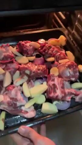 A hand slides a metal baking tray into an oven. The tray is filled with raw meat chunks, potato wedges, and quartered red onions. The oven is dark inside.