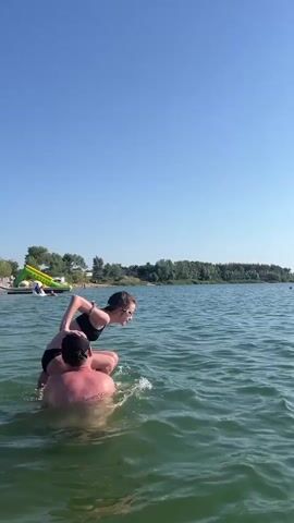A man holds a woman on his shoulders in the water, and she jumps off him into the lake. He then dives in after her. The scene takes place on a clear, sunny day with a blue sky and a treelined shore in the distance.