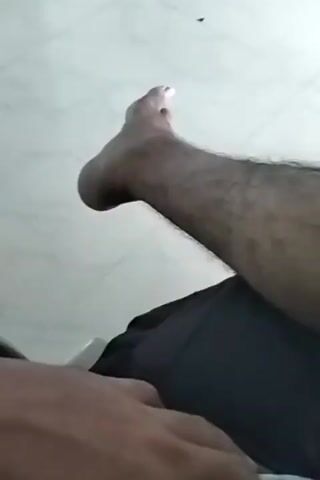 The video shows a close-up view of a person's legs and feet. The person's legs are hairy and they are wearing black shorts. A patterned cloth is visible under their legs. The video appears to be filmed indoors, as indicated by the tiled floor in the background. The camera angle is low, suggesting it might be held by the person themselves. The lighting is natural and seems to be coming from overhead. The overall impression is a casual, home-like environment.