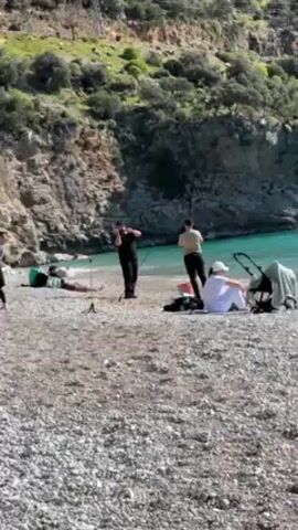 The video shows a pebbled beach with a rocky, vegetated hillside in the background. A man in black clothing stands near a microphone stand, seemingly talking or performing. A woman in a tan top and black pants interacts with him, possibly filming or directing. A person in white sits on the beach with a stroller nearby. Another individual lies on a towel in the background. The turquoise water of the bay can be seen lapping at the shore. The sky is sunny, suggesting a pleasant day at the beach.