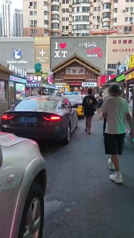 The video shows a street scene with traffic and pedestrians. In the foreground, a dark-colored Audi car is visible with its taillights illuminated. To the right, several people are walking and standing. In the background, there are buildings with various signs, including "luckin coffee" and what appears to be a "I LOVE XT" sign with Korean text. The street is lined with small shops or food stalls. The overall atmosphere suggests a busy urban environment, possibly a commercial district.