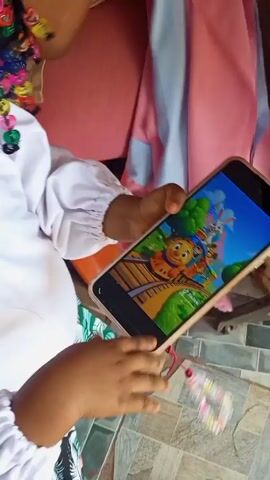 A young child is looking at a phone and watching a cartoon. The child has dark braided hair and is wearing a white long-sleeved shirt. The phone screen displays an animated cartoon featuring a train and other characters. The child is sitting on a person's lap and there is an orange plastic chair nearby. The child smiles, looks at the phone, and laughs. The camera focuses on the child's hair, showing the braids and colorful hair ties.