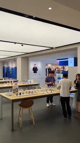 This video shows the interior of a modern retail store, possibly an electronics store, with a bright, white, illuminated ceiling and dark flooring. Several long display tables are set up throughout the space, each showcasing various products, likely mobile phones or smartwatches, arranged neatly. A few customers and employees are present in the store. In the foreground, a man in a blue shirt and black pants stands near a display table, appearing to examine a product. Another man in a white shirt and black pants walks away from the camera. Further into the store, more customers are seen interacting with the displays. On the walls, large digital screens display product imagery. The overall atmosphere suggests a clean, organized, and contemporary retail environment.