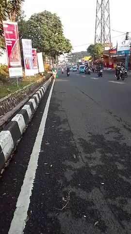 The video shows a street in an urban environment, with traffic flowing in both directions. Cars, motorcycles, and people are visible. There are trees, buildings, and advertising billboards on either side of the road. A large electricity pylon stands tall in the background. The road is paved with asphalt, and there is a curb separating the road from a pedestrian area. The video is shot from a stationary position, offering a wide view of the street.