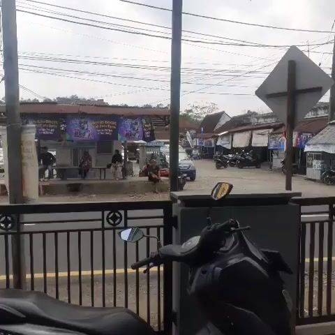 The video shows a street scene with a black scooter in the foreground. In the background, there are several buildings that appear to be shops or stalls. There are people walking around and sitting on benches. There are also several motorcycles parked. The sky is cloudy. There are banners hanging from one of the buildings. A car is driving past. The video is shaky and appears to have been taken from a moving vehicle.