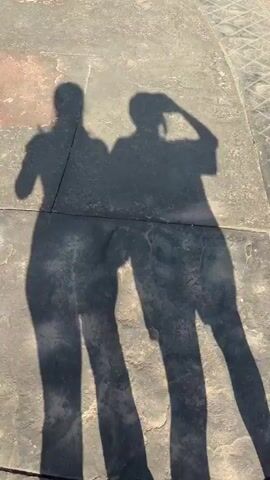The video displays two dark, distinct shadows of people cast on a light-colored, textured ground, appearing to be concrete or pavement. The shadow on the left depicts a person standing still, with long hair or a head covering. The shadow on the right shows another person standing, with one arm raised to their head, as if drinking or adjusting headwear. Both shadows remain static throughout the brief clip. In the bottom right corner, a human foot wearing a light blue sandal with pink painted toenails is visible. The scene is illuminated by bright sunlight, creating sharp shadow edges.