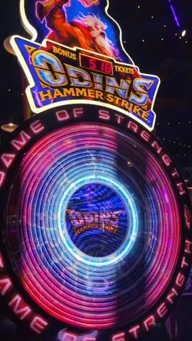 The video begins with a close-up of a large, illuminated arcade game called "ODIN'S HAMMER STRIKE." The game features a spinning circular display with neon lights in pink, blue, and purple. The game's title and an image of Odin are visible at the top. The camera then pans out to reveal a dimly lit arcade with various other games and a large overhead fan. Several people are visible in the background, some appearing to be playing games. The video ends with a shot of the arcade's ceiling, with lights and a large industrial fan.