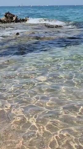 The video shows a close-up view of clear water lapping at a rocky shore. The water is transparent, revealing a sandy bottom and the texture of small rocks submerged beneath the surface. Sunlight creates dappled patterns on the water, indicating a bright, sunny day. In the background, the ocean stretches to the horizon under a clear blue sky, with a few distant sailboats or buoys visible. The waves gently break against the rocks, creating a sense of calm and natural beauty.