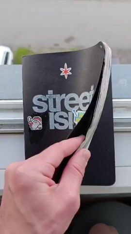 A person is holding a black-covered book with their left hand, rapidly flipping through its pages. The book is open, and various colorful drawings and scribbles are visible on the pages as they turn. The person's thumb and fingers are positioned on the edge of the pages, facilitating the quick flipping motion. The background shows an outdoor setting with what appears to be a light-colored building or wall and a glimpse of a vehicle.