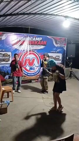 Three people are on a paved outdoor area under a roof. A banner behind them reads "TOUR CAMP ADEKAN". The person on the left, a male wearing a red t-shirt and jeans, stands facing forward. The person in the middle, a male wearing a black t-shirt, khaki pants, and a beanie, holds a microphone. The person on the right, a female wearing a black t-shirt, a black skirt, and a light blue beanie, holds a microphone and appears to be dancing or performing. There are chairs and boxes in the foreground. A speaker is visible on the right side of the frame.