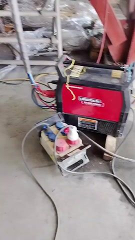The video displays an industrial setting, likely a workshop or construction area. The primary focus is a red and black Lincoln Electric Invertec welder, positioned on a concrete floor. Connected to the welder are various cables, including red, blue, and yellow ones. Beside the welder is a power distribution box, featuring multiple blue and red industrial-style plugs and sockets, indicating connections for different power phases. A metal rod is partially visible in the foreground, and wooden blocks are used under the welder. In the background, there are metal structures and possibly some wrapped items, further suggesting an industrial environment. No people are visible, but the setup implies preparations for or an ongoing welding operation. The camera slowly pans around the equipment.