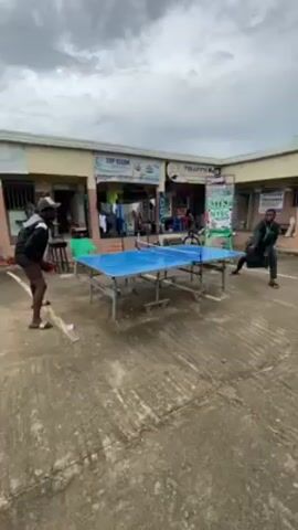 Two individuals engage in a table tennis match on an outdoor, blue-surfaced table. One player, on the left, is attired in shorts, a black jacket, a hat, and sandals. The second player, on the right, wears a green robe. The game transpires on a concrete surface, with a backdrop consisting of a row of modest, single-story buildings under a cloudy sky. The buildings are adorned with signage, including "TOP CLEAN" and "TURAFI'S".
