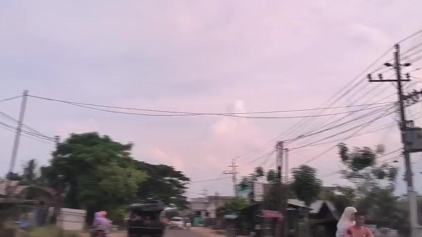 The video depicts a street scene in what appears to be a residential area. The shot is taken from a moving vehicle, looking forward along the road. Several vehicles are visible, including a truck carrying passengers in the back, a white minivan, and a person riding a motorcycle. The motorcycle rider is wearing a purple headscarf. Buildings and homes line the street, interspersed with trees and utility poles with overhead wires. The sky is overcast with pale clouds.