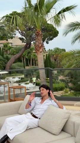 A young man in a white shirt and white pants is sitting on a light-colored couch with his arms spread wide. He is smiling and looking towards the camera. He has dark hair and is wearing a brown belt. In the background, there are palm trees, lush greenery, and some buildings. There are also tables with white tablecloths and umbrellas visible, suggesting an outdoor dining area. The man appears to be on a terrace or balcony overlooking a landscaped area.