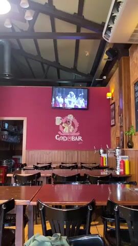 The video shows the interior of a bar. There are several tables with dark chairs arranged in rows. The walls are painted a deep red and decorated with a mural that appears to be a character from a video game holding a beer mug, with the words "GOD OF BAR" written below. A flat-screen television is mounted on the wall above the mural, displaying a variety of video content, including music videos and advertisements. The ceiling has exposed wooden beams and lighting fixtures. The overall atmosphere suggests a casual dining or drinking establishment.