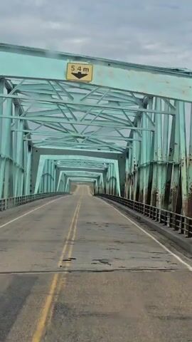 The video shows a forward-facing view from a vehicle driving onto a long, green steel truss bridge. The road surface is asphalt with yellow dividing lines and appears worn with some potholes. The bridge has a repetitive, arched structure overhead. A sign with a yellow background and black text indicating a height limit of 3.4 meters is visible at the beginning of the bridge. The sky is overcast. There are no people or other vehicles visible on the bridge. The camera is positioned as if it is the driver's point of view.