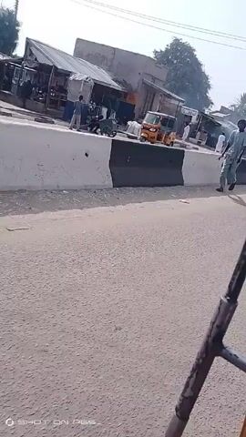The video shows a man running across a road in what appears to be a market or town setting. The man is wearing light colored clothing and a dark hat. He is moving from left to right across the frame, away from a barrier dividing the road. In the background are buildings and vehicles, including a yellow three-wheeled vehicle. Other people are visible in the background, some standing near the buildings. The road is paved and the weather appears to be sunny. A portion of what may be a vehicle is visible in the bottom right of the frame. The lower portion of the frame has "SHOT ON PES" displayed.